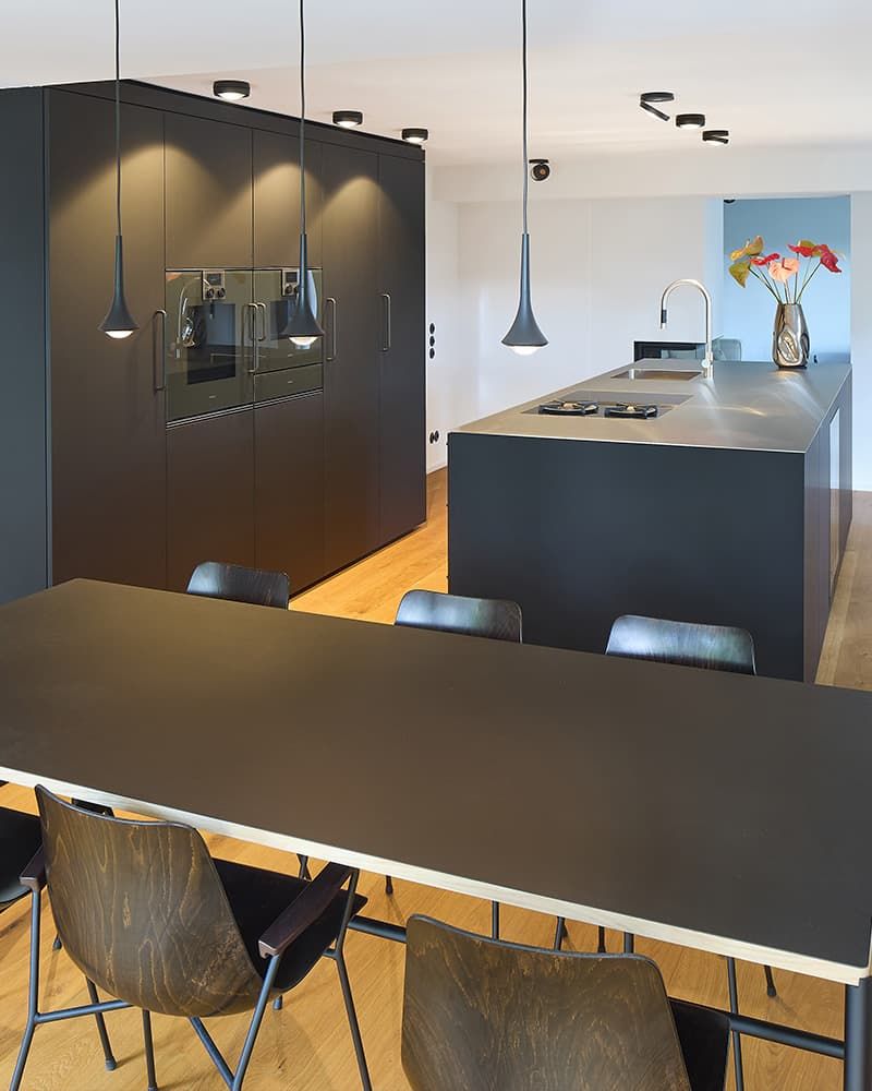 Eine edle schwarze Küche aus Holz, mit großem Wandschrank und eingebauten Ofen. Freistehende Kücheninsel mit Waschbecken und Kochfeldern, sowie ein großer Esstisch im Vordergrund.