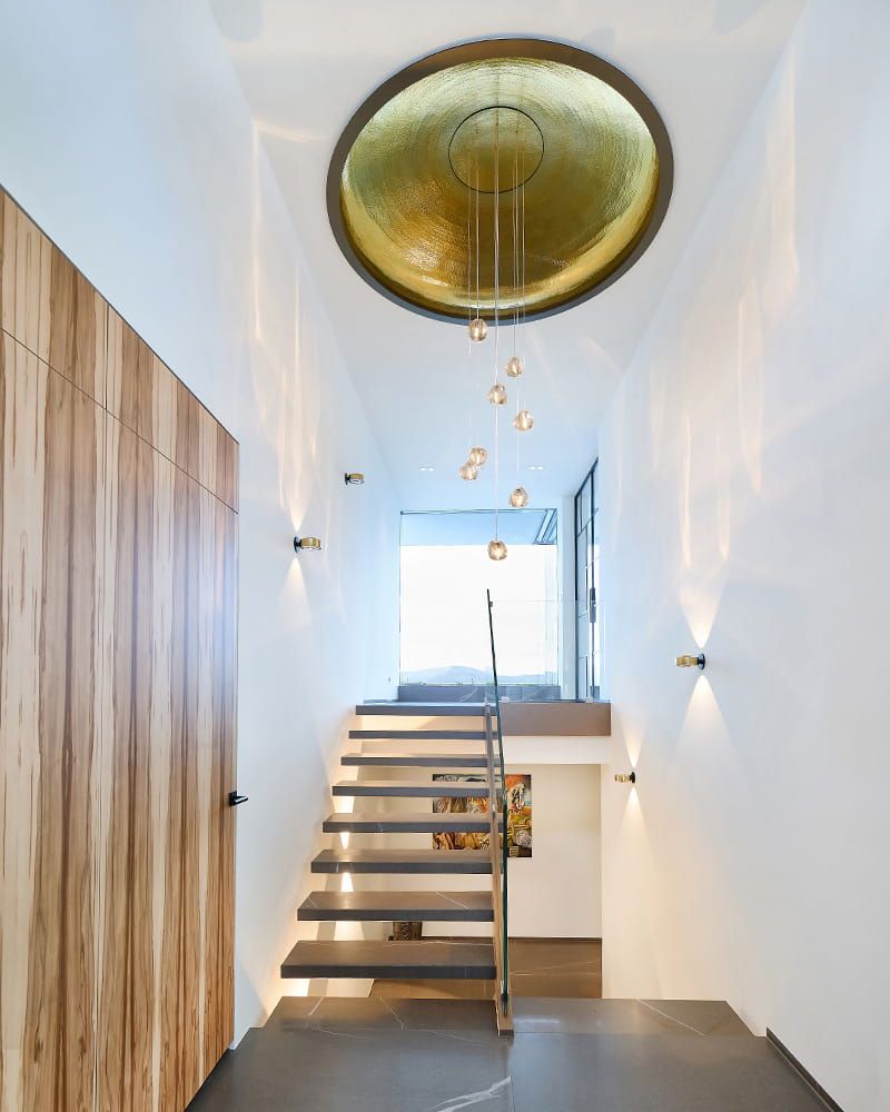 Ein stillvoller Eingang mit schwarzen Boden, einer großen Regalseite auf der linken Seite. In der Mitte ist eine Treppe nach oben und unten, mit goldenem Deckenelement.