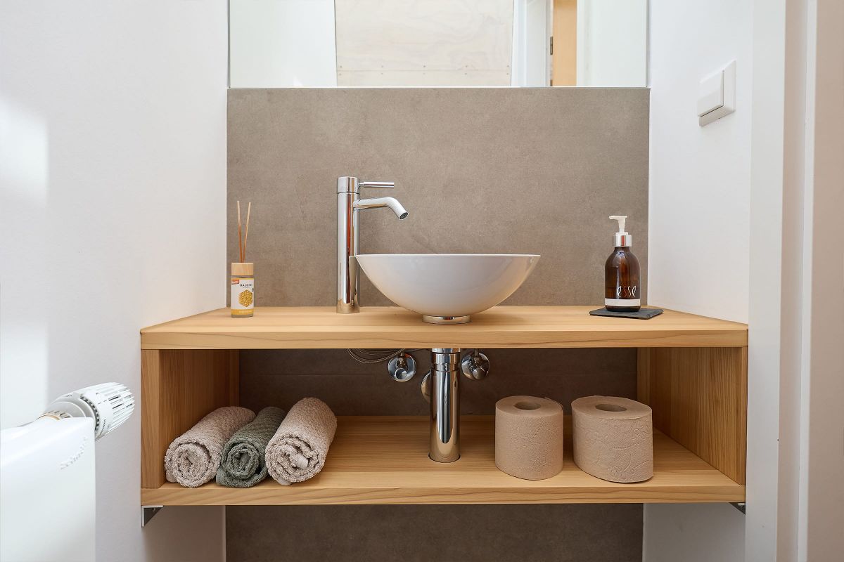 Kleines Waschbecken mit Holzregal, grauer Wandverkleidung und minimalistischen Accessoires in einem modernen Bad.