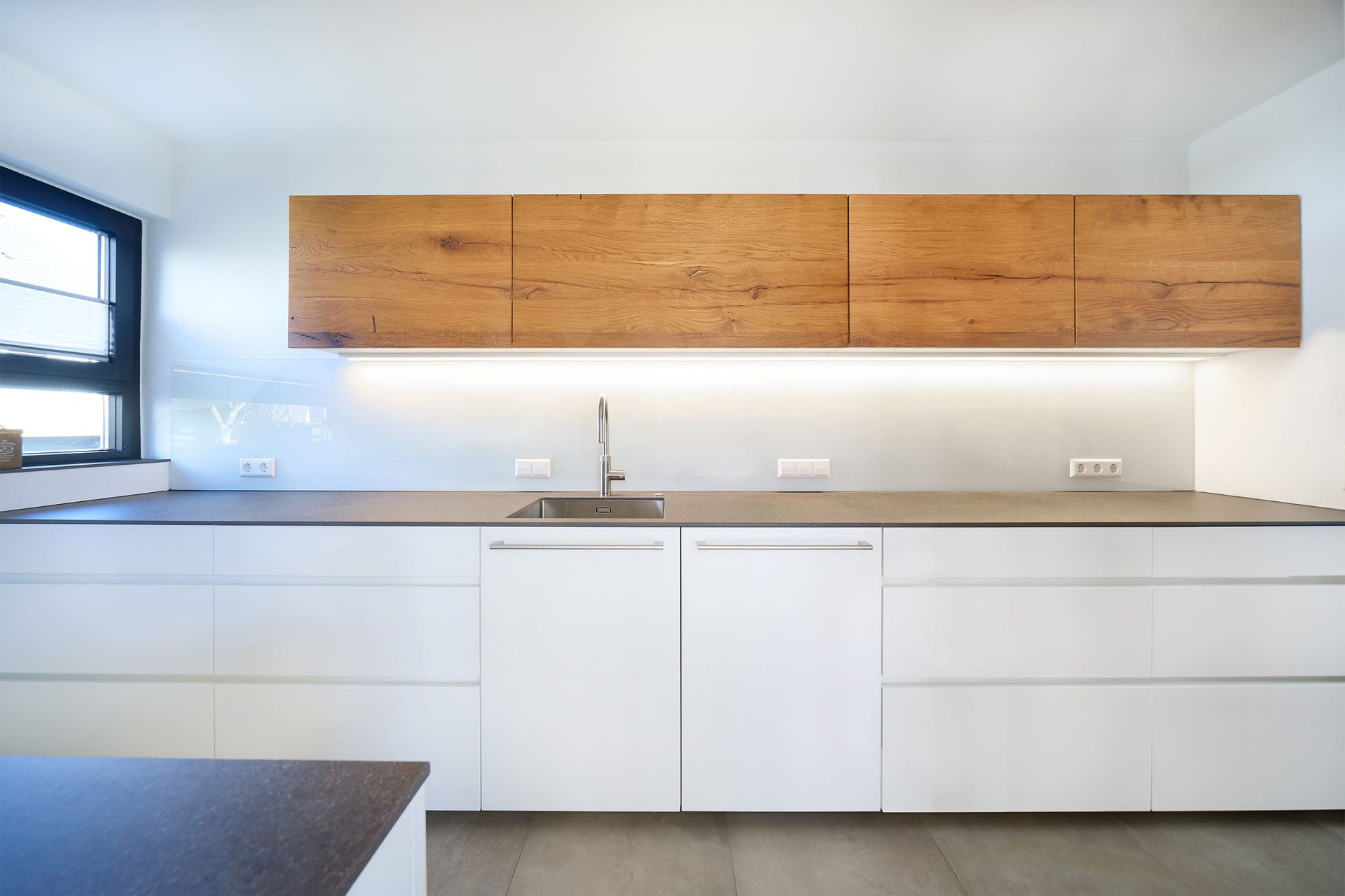 Moderne Küche mit weißen Fronten, dunkler Arbeitsplatte und Oberschränken aus warmem Holz in klarem, puristischem Design.